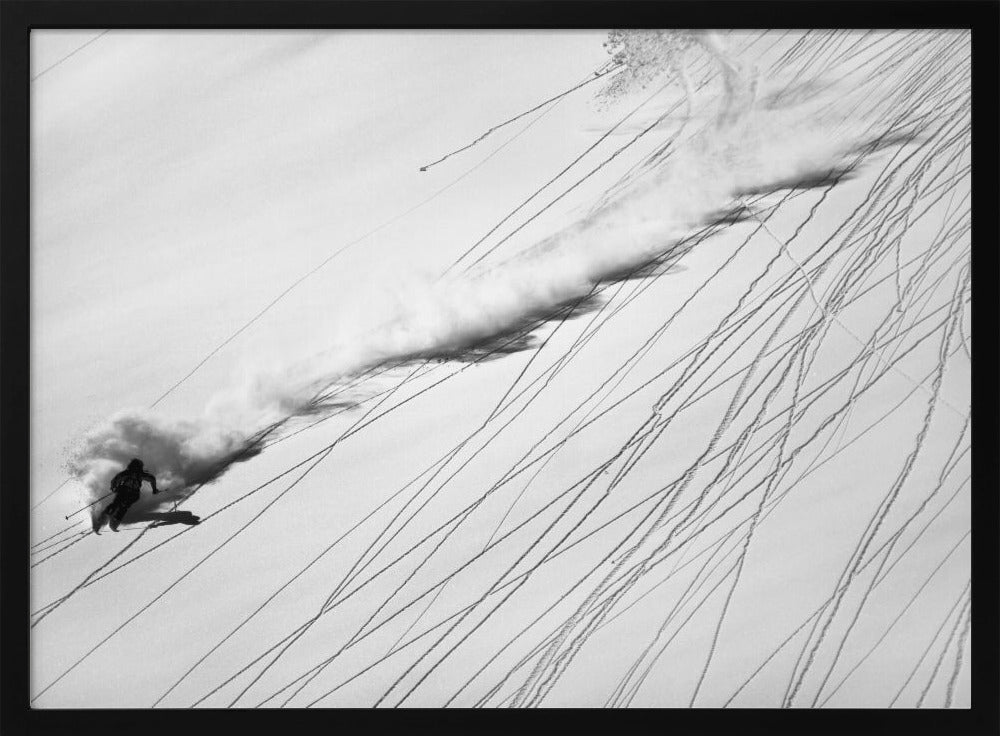 A dramatic black and white overhead photograph of a skier carving down a steep, powder-filled mountain slope, kicking up a huge plume of snow. Numerous ski tracks crisscross the pristine white snow, creating a pattern of dark lines. Poster