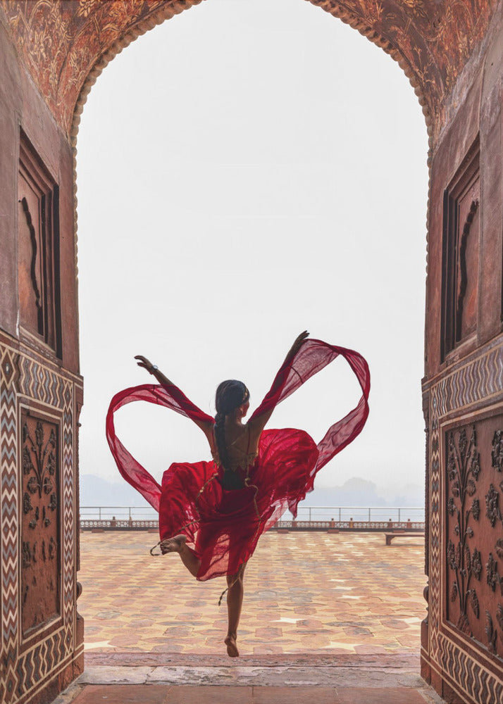 A woman in a flowing red dress dances joyfully, framed by an ornate, carved archway. Seen from behind, she leaps with her arms outstretched, holding a long red scarf that forms a heart shape around her against a bright, hazy sky. Poster
