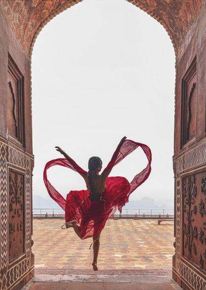 A woman in a flowing red dress dances joyfully, framed by an ornate, carved archway. Seen from behind, she leaps with her arms outstretched, holding a long red scarf that forms a heart shape around her against a bright, hazy sky. Poster