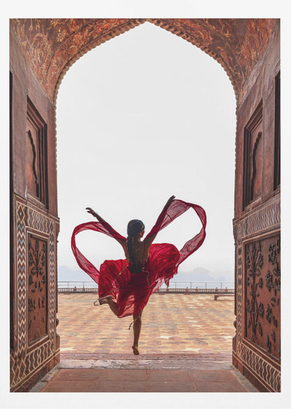 A woman in a flowing red dress dances joyfully, framed by an ornate, carved archway. Seen from behind, she leaps with her arms outstretched, holding a long red scarf that forms a heart shape around her against a bright, hazy sky. Poster