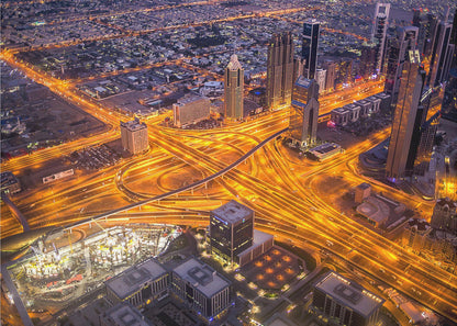 A stunning aerial view of a futuristic city at night, with brilliantly lit highways forming intricate patterns of golden light amidst towering skyscrapers, enclosed in a silver frame. Poster