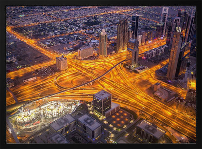 A stunning aerial view of a futuristic city at night, with brilliantly lit highways forming intricate patterns of golden light amidst towering skyscrapers, enclosed in a silver frame. Poster