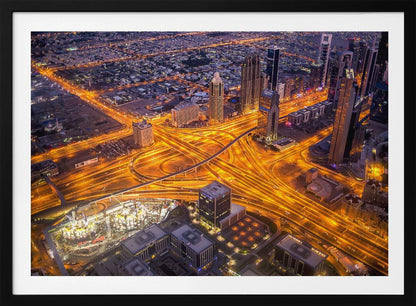 A stunning aerial view of a futuristic city at night, with brilliantly lit highways forming intricate patterns of golden light amidst towering skyscrapers, enclosed in a silver frame. Poster