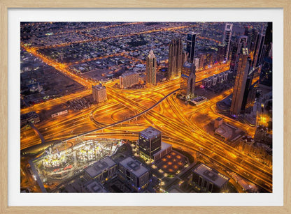 A stunning aerial view of a futuristic city at night, with brilliantly lit highways forming intricate patterns of golden light amidst towering skyscrapers, enclosed in a silver frame. Poster