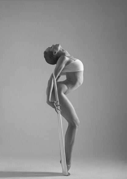 A framed black and white photograph of a graceful female dancer arching her back dramatically. She holds a long white elastic band that stretches down her body to her foot, emphasizing her athletic form and flexibility against a plain studio background. Print