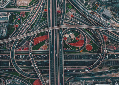 An aerial, top-down photograph of a sprawling and complex highway interchange, framed in silver. Multiple levels of roads and overpasses curve and intertwine, with blurred streaks of traffic visible. The spaces between the highways feature manicured green lawns and artistic red and patterned landscaping. Wall Art