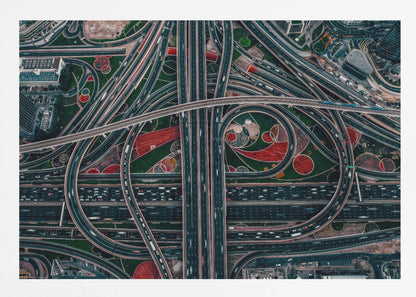 An aerial, top-down photograph of a sprawling and complex highway interchange, framed in silver. Multiple levels of roads and overpasses curve and intertwine, with blurred streaks of traffic visible. The spaces between the highways feature manicured green lawns and artistic red and patterned landscaping. Wall Art