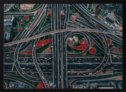 An aerial, top-down photograph of a sprawling and complex highway interchange, framed in silver. Multiple levels of roads and overpasses curve and intertwine, with blurred streaks of traffic visible. The spaces between the highways feature manicured green lawns and artistic red and patterned landscaping. Wall Art