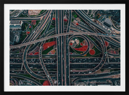 An aerial, top-down photograph of a sprawling and complex highway interchange, framed in silver. Multiple levels of roads and overpasses curve and intertwine, with blurred streaks of traffic visible. The spaces between the highways feature manicured green lawns and artistic red and patterned landscaping. Wall Art