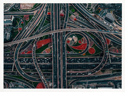 An aerial, top-down photograph of a sprawling and complex highway interchange, framed in silver. Multiple levels of roads and overpasses curve and intertwine, with blurred streaks of traffic visible. The spaces between the highways feature manicured green lawns and artistic red and patterned landscaping. Wall Art