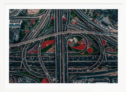 An aerial, top-down photograph of a sprawling and complex highway interchange, framed in silver. Multiple levels of roads and overpasses curve and intertwine, with blurred streaks of traffic visible. The spaces between the highways feature manicured green lawns and artistic red and patterned landscaping. Wall Art