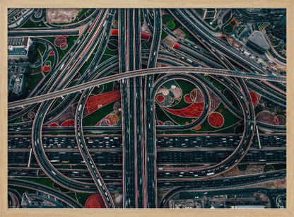 An aerial, top-down photograph of a sprawling and complex highway interchange, framed in silver. Multiple levels of roads and overpasses curve and intertwine, with blurred streaks of traffic visible. The spaces between the highways feature manicured green lawns and artistic red and patterned landscaping. Wall Art