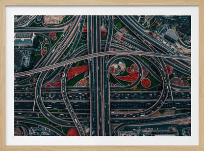 An aerial, top-down photograph of a sprawling and complex highway interchange, framed in silver. Multiple levels of roads and overpasses curve and intertwine, with blurred streaks of traffic visible. The spaces between the highways feature manicured green lawns and artistic red and patterned landscaping. Wall Art