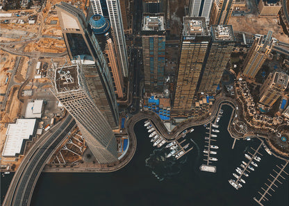 An aerial, high-angle photograph looking down on a modern city skyline, featuring towering skyscrapers, including a distinctive twisted tower, clustered around a dark-water marina filled with white yachts. A multi-lane highway snakes through the urban landscape, which has a warm, desaturated color palette and is enclosed in a silver frame. Decor