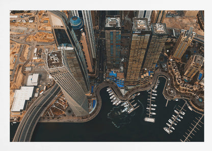 An aerial, high-angle photograph looking down on a modern city skyline, featuring towering skyscrapers, including a distinctive twisted tower, clustered around a dark-water marina filled with white yachts. A multi-lane highway snakes through the urban landscape, which has a warm, desaturated color palette and is enclosed in a silver frame. Decor