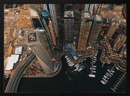 An aerial, high-angle photograph looking down on a modern city skyline, featuring towering skyscrapers, including a distinctive twisted tower, clustered around a dark-water marina filled with white yachts. A multi-lane highway snakes through the urban landscape, which has a warm, desaturated color palette and is enclosed in a silver frame. Decor