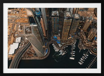 An aerial, high-angle photograph looking down on a modern city skyline, featuring towering skyscrapers, including a distinctive twisted tower, clustered around a dark-water marina filled with white yachts. A multi-lane highway snakes through the urban landscape, which has a warm, desaturated color palette and is enclosed in a silver frame. Decor