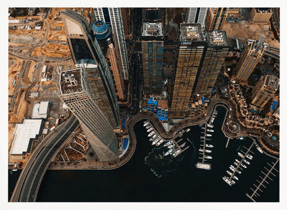An aerial, high-angle photograph looking down on a modern city skyline, featuring towering skyscrapers, including a distinctive twisted tower, clustered around a dark-water marina filled with white yachts. A multi-lane highway snakes through the urban landscape, which has a warm, desaturated color palette and is enclosed in a silver frame. Decor