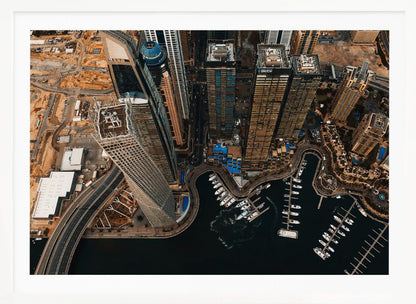 An aerial, high-angle photograph looking down on a modern city skyline, featuring towering skyscrapers, including a distinctive twisted tower, clustered around a dark-water marina filled with white yachts. A multi-lane highway snakes through the urban landscape, which has a warm, desaturated color palette and is enclosed in a silver frame. Decor