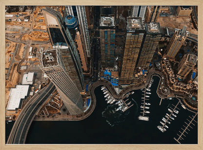 An aerial, high-angle photograph looking down on a modern city skyline, featuring towering skyscrapers, including a distinctive twisted tower, clustered around a dark-water marina filled with white yachts. A multi-lane highway snakes through the urban landscape, which has a warm, desaturated color palette and is enclosed in a silver frame. Decor