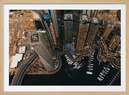 An aerial, high-angle photograph looking down on a modern city skyline, featuring towering skyscrapers, including a distinctive twisted tower, clustered around a dark-water marina filled with white yachts. A multi-lane highway snakes through the urban landscape, which has a warm, desaturated color palette and is enclosed in a silver frame. Decor