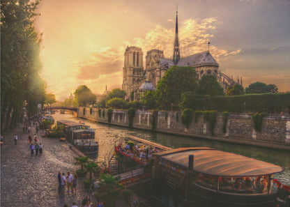 A scenic photograph of the Notre Dame Cathedral in Paris during a warm, golden sunset, as viewed from across the River Seine. People are strolling along the cobblestone riverbank and riding on tour boats docked on the water. The image is presented within a silver picture frame. Decor