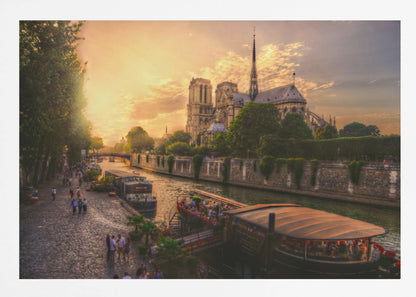 A scenic photograph of the Notre Dame Cathedral in Paris during a warm, golden sunset, as viewed from across the River Seine. People are strolling along the cobblestone riverbank and riding on tour boats docked on the water. The image is presented within a silver picture frame. Decor