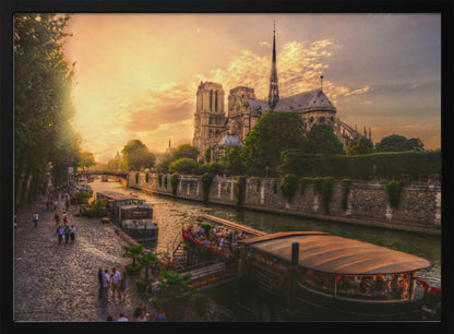 A scenic photograph of the Notre Dame Cathedral in Paris during a warm, golden sunset, as viewed from across the River Seine. People are strolling along the cobblestone riverbank and riding on tour boats docked on the water. The image is presented within a silver picture frame. Decor