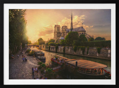 A scenic photograph of the Notre Dame Cathedral in Paris during a warm, golden sunset, as viewed from across the River Seine. People are strolling along the cobblestone riverbank and riding on tour boats docked on the water. The image is presented within a silver picture frame. Decor