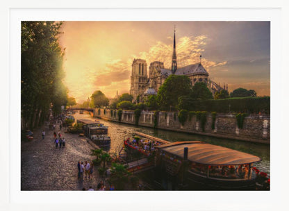 A scenic photograph of the Notre Dame Cathedral in Paris during a warm, golden sunset, as viewed from across the River Seine. People are strolling along the cobblestone riverbank and riding on tour boats docked on the water. The image is presented within a silver picture frame. Decor