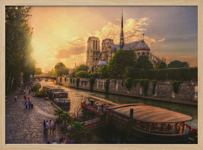 A scenic photograph of the Notre Dame Cathedral in Paris during a warm, golden sunset, as viewed from across the River Seine. People are strolling along the cobblestone riverbank and riding on tour boats docked on the water. The image is presented within a silver picture frame. Decor