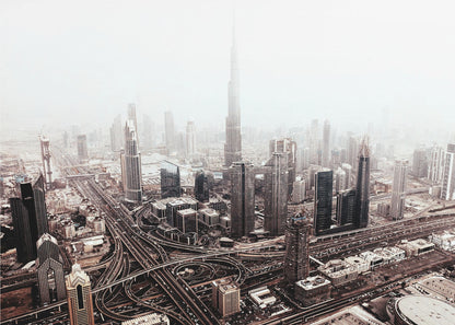 An aerial photograph of a sprawling modern cityscape on a hazy day, framed in silver. The iconic Burj Khalifa stands tall in the center background, surrounded by numerous other skyscrapers and a complex network of highways and interchanges in the foreground, all rendered in a muted, slightly sepia-toned color palette. Print