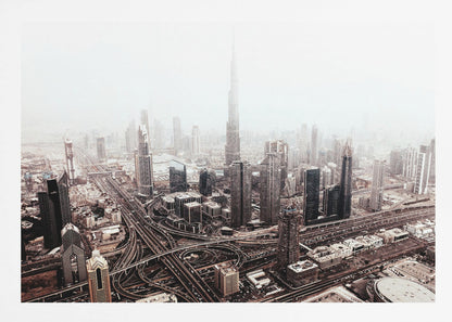 An aerial photograph of a sprawling modern cityscape on a hazy day, framed in silver. The iconic Burj Khalifa stands tall in the center background, surrounded by numerous other skyscrapers and a complex network of highways and interchanges in the foreground, all rendered in a muted, slightly sepia-toned color palette. Print