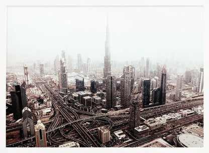 An aerial photograph of a sprawling modern cityscape on a hazy day, framed in silver. The iconic Burj Khalifa stands tall in the center background, surrounded by numerous other skyscrapers and a complex network of highways and interchanges in the foreground, all rendered in a muted, slightly sepia-toned color palette. Print