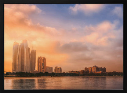 A framed photograph of the Abu Dhabi city skyline at sunset, featuring modern skyscrapers and the Emirates Palace hotel glowing in warm, orange light. The buildings are reflected in the calm water of the foreground, under a sky with soft orange and blue clouds. Poster