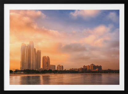 A framed photograph of the Abu Dhabi city skyline at sunset, featuring modern skyscrapers and the Emirates Palace hotel glowing in warm, orange light. The buildings are reflected in the calm water of the foreground, under a sky with soft orange and blue clouds. Poster
