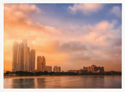 A framed photograph of the Abu Dhabi city skyline at sunset, featuring modern skyscrapers and the Emirates Palace hotel glowing in warm, orange light. The buildings are reflected in the calm water of the foreground, under a sky with soft orange and blue clouds. Poster
