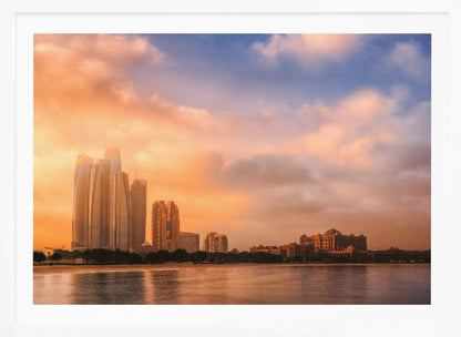A framed photograph of the Abu Dhabi city skyline at sunset, featuring modern skyscrapers and the Emirates Palace hotel glowing in warm, orange light. The buildings are reflected in the calm water of the foreground, under a sky with soft orange and blue clouds. Poster