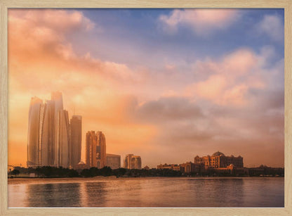 A framed photograph of the Abu Dhabi city skyline at sunset, featuring modern skyscrapers and the Emirates Palace hotel glowing in warm, orange light. The buildings are reflected in the calm water of the foreground, under a sky with soft orange and blue clouds. Poster