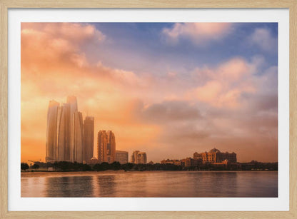 A framed photograph of the Abu Dhabi city skyline at sunset, featuring modern skyscrapers and the Emirates Palace hotel glowing in warm, orange light. The buildings are reflected in the calm water of the foreground, under a sky with soft orange and blue clouds. Poster