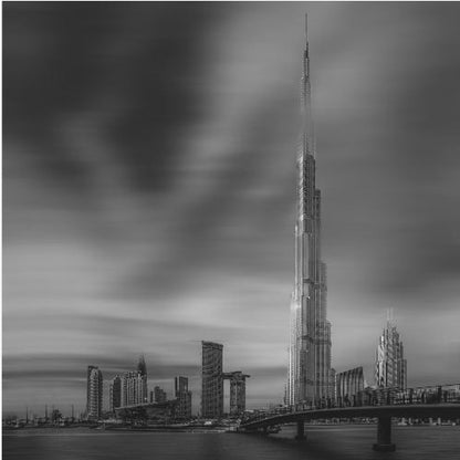 A framed, long-exposure black and white photograph of the Dubai skyline, featuring the iconic Burj Khalifa. The sky has a dramatic, streaky appearance from the long exposure, contrasting with the sharp details of the modern architecture. A bridge crosses the water in the foreground. Print