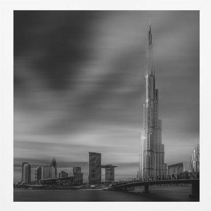 A framed, long-exposure black and white photograph of the Dubai skyline, featuring the iconic Burj Khalifa. The sky has a dramatic, streaky appearance from the long exposure, contrasting with the sharp details of the modern architecture. A bridge crosses the water in the foreground. Print