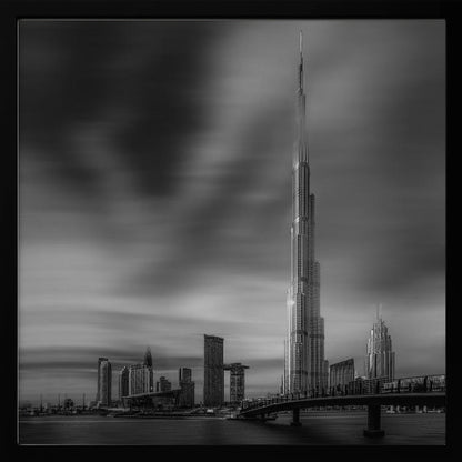 A framed, long-exposure black and white photograph of the Dubai skyline, featuring the iconic Burj Khalifa. The sky has a dramatic, streaky appearance from the long exposure, contrasting with the sharp details of the modern architecture. A bridge crosses the water in the foreground. Print