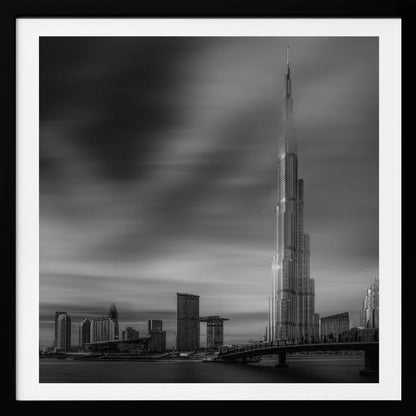 A framed, long-exposure black and white photograph of the Dubai skyline, featuring the iconic Burj Khalifa. The sky has a dramatic, streaky appearance from the long exposure, contrasting with the sharp details of the modern architecture. A bridge crosses the water in the foreground. Print