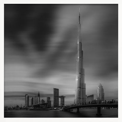 A framed, long-exposure black and white photograph of the Dubai skyline, featuring the iconic Burj Khalifa. The sky has a dramatic, streaky appearance from the long exposure, contrasting with the sharp details of the modern architecture. A bridge crosses the water in the foreground. Print