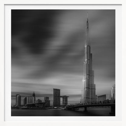 A framed, long-exposure black and white photograph of the Dubai skyline, featuring the iconic Burj Khalifa. The sky has a dramatic, streaky appearance from the long exposure, contrasting with the sharp details of the modern architecture. A bridge crosses the water in the foreground. Print