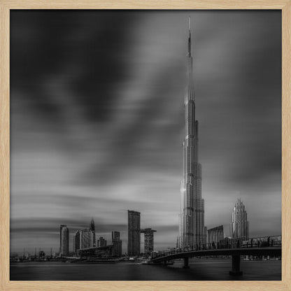 A framed, long-exposure black and white photograph of the Dubai skyline, featuring the iconic Burj Khalifa. The sky has a dramatic, streaky appearance from the long exposure, contrasting with the sharp details of the modern architecture. A bridge crosses the water in the foreground. Print
