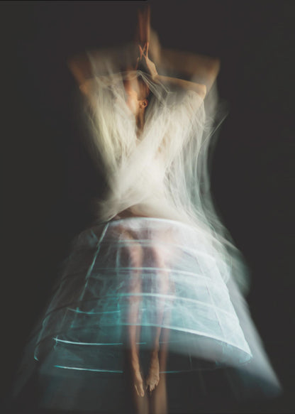 An ethereal, long-exposure photograph of a dancing figure against a dark gray background. The motion blur creates a ghostly effect, with white, gauzy fabric swirling around the upper body. The figure wears a transparent crinoline hoop skirt, revealing her legs and feet. Artwork