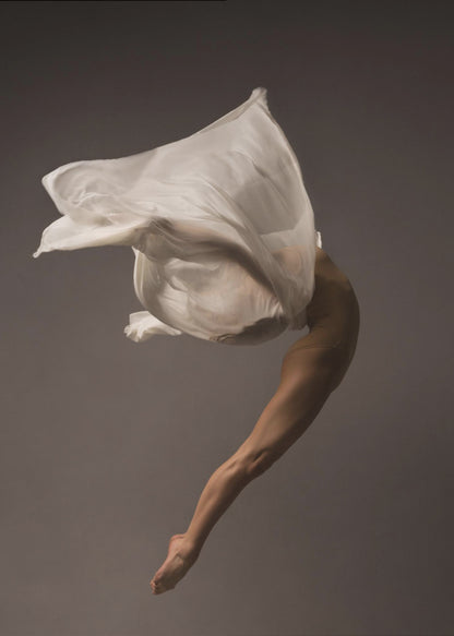 A minimalist fine art photograph of a dancer in mid-air against a solid grey background. The dancer's body is arched backwards, with a large piece of flowing white fabric enveloping their torso and creating a sense of dynamic, weightless motion. Only their muscular leg and pointed foot are clearly visible, emphasizing grace and form. Wall Art