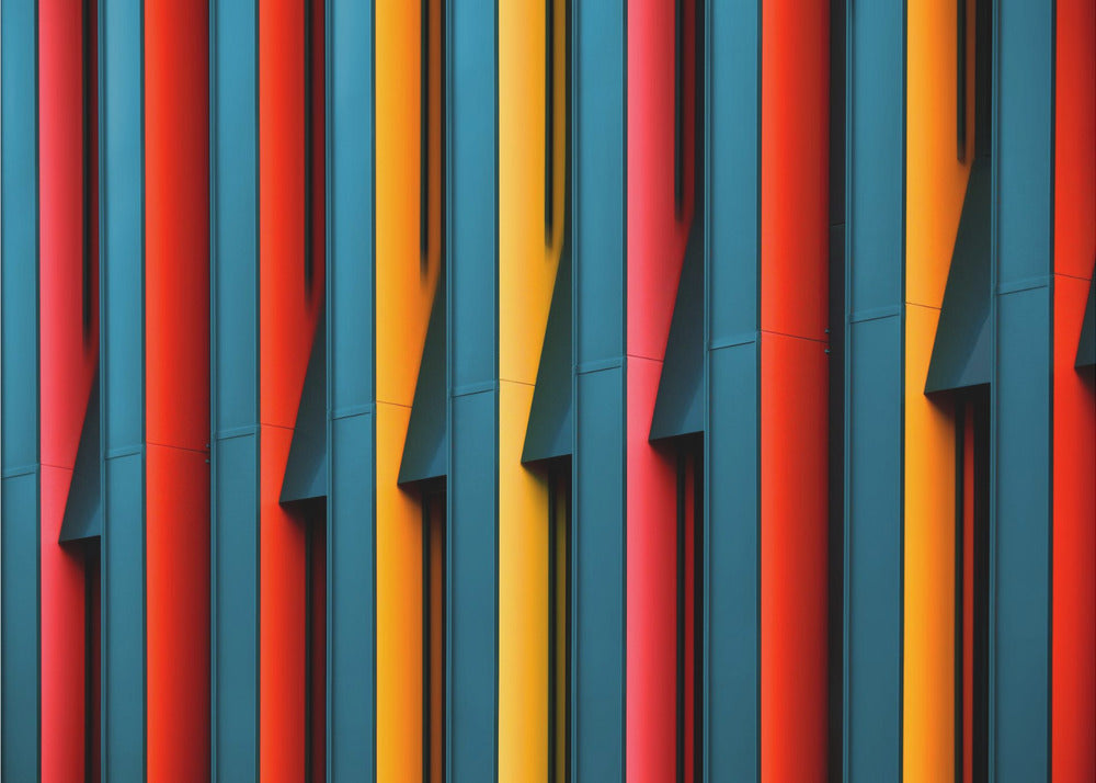 A close-up abstract photograph of a colorful modern building facade, featuring a repeating pattern of vertical fins in vibrant red, teal blue, and yellow-orange, creating a dynamic interplay of light, shadow, and geometric lines, all enclosed in a silver frame. Artwork